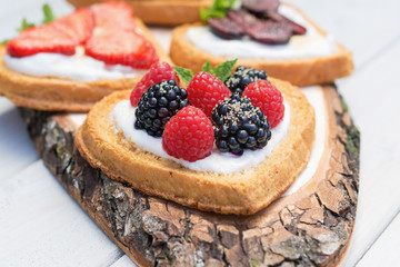 Heart shaped biscuits spread with quark, strawberries, blackberries, raspberries, cherries and a twig of mint presented on a tree disk