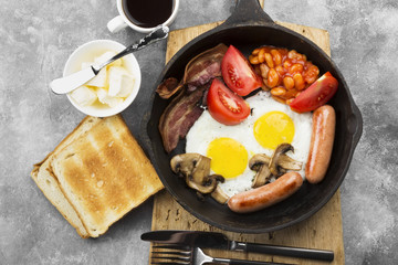 Traditional English breakfast - bacon, sausages, fried eggs, tomatoes, beans, mushrooms, toast with butter -  in cooking pan and coffee on a gray background. Top view. Food background