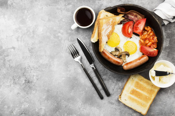 Traditional English breakfast - bacon, sausages, fried eggs, tomatoes, beans, mushrooms, toast with butter -  in cooking pan and coffee on a gray background. Top view, copy space. Food background