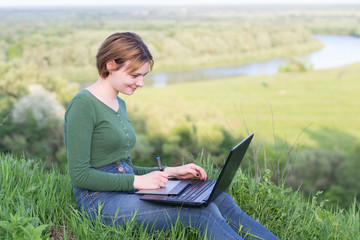 Beautiful young girl using her graphic tablet sitting in the grass near river