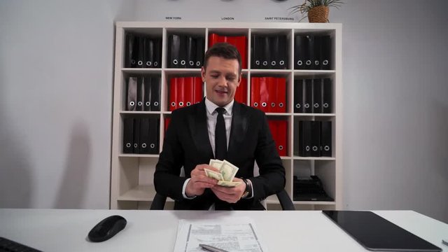 4k uhd POV Young attractive business man in black suit and tie counting dollars with smile. The promise that the work and servise will be done. Hide money in pocket. Office indoor.