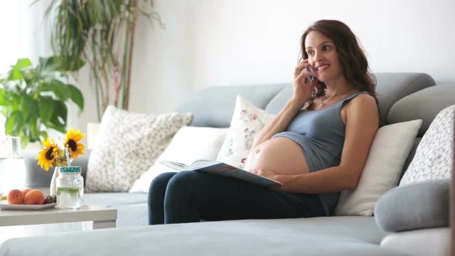 Young pretty pregnant woman speaking on the phone at home on the couch, eating healthy, fruits, lemonade and flowers on the table