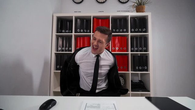 4k POV. Young attractive Handsome businessman funny dancing in office armchair necktie. He strip remove his black suit. Computer, mouse, keyboard, digital pad, shelf with folders. Wide shot.