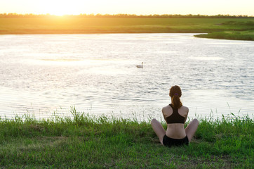 Naklejka na ściany i meble Young woman meditating in lotus position on the river bank, sunset. A woman with a healthy lifestyle, a copy of the free space.