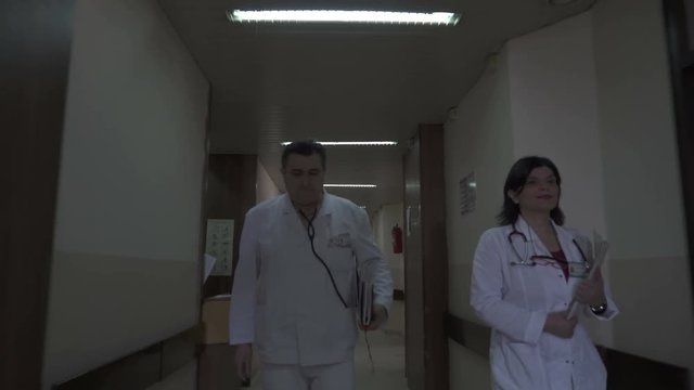 Mature Male And Middle Age Female Doctors Holding Documentation, Walking Through The Hospital Darker Hallway, Entering The Patient's Room To Performing Examination Of Very Old Woman, Steady Cam