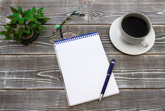 Notebook paper clean sheet, blue pen, glasses, coffee cup, decorative plant on the background table wood texture. Ready for inserting text or mockup.