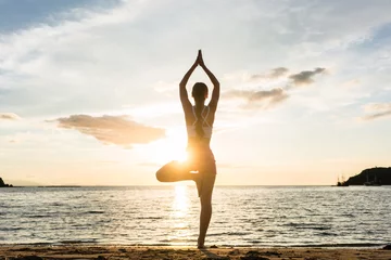 Abwaschbare Fototapete Yogaschule Rückansicht der Silhouette einer Frau in voller Länge, die auf einem Bein steht, während sie die Baum-Yoga-Pose an einem ruhigen Strand praktiziert, aufgenommen bei Sonnenuntergang während der Sommerferien in Indonesien