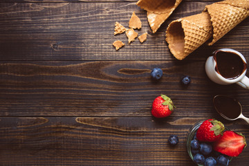 fresh berries, waffle cone and chocolate sauce on the wooden background, top view. Ice-cream background with copy space