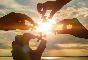 Collaborate four hands trying to connect a puzzle piece with a sunset background. A puzzle in hand against sunlight. One part of the whole. Symbol of association and communication. Business strategy.