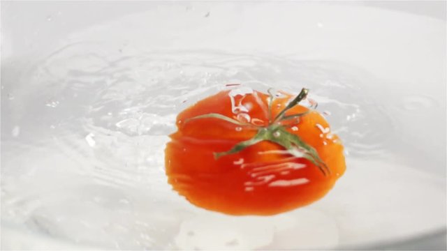 Slow motion. One red ripe tomato with green leaves falls under water super slow motion video. Splash and spray. White background. High-speed-camera phantom flex.