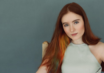 Cropped portrait of the beautiful female model with red long hair and gorgeous green eyes against blue wall in studio. Attractive young woman looking at camera with thoughtful and dreamy expression