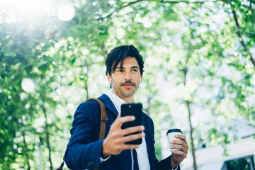 Fototapeta premium Stylish young businessman using smartphone for listining music while walking in city park and holding take away coffee hand.Horizontal,blurred background.