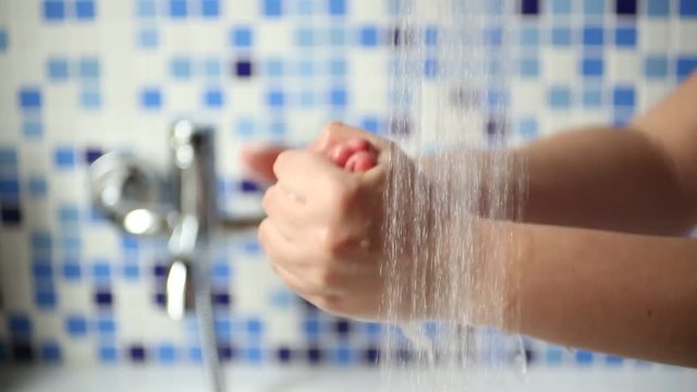 A young woman is washing hands under the water falling from the shower faucet in a bathroom. The wall is gres tiled with blue and white tiles.