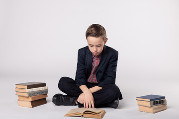 Cute boy in a suit sitting and and reading a book on a white background. pile of books.