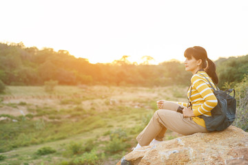Young Woman in yellow sweater’s sitting on a giant rock for rest and watching sunset between travel in mountain for her vacation.