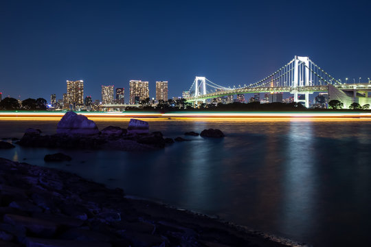 Rainbow Bridge located in Tokyo Bay at night seen from Odaiba shore light traces of the passing boats are reflecting in the water
