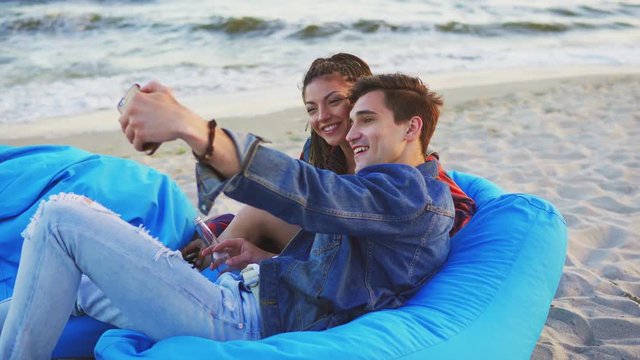 An couple having fun and taking selfies laying on easy chairs with blanket on the beach. Shot in 4k