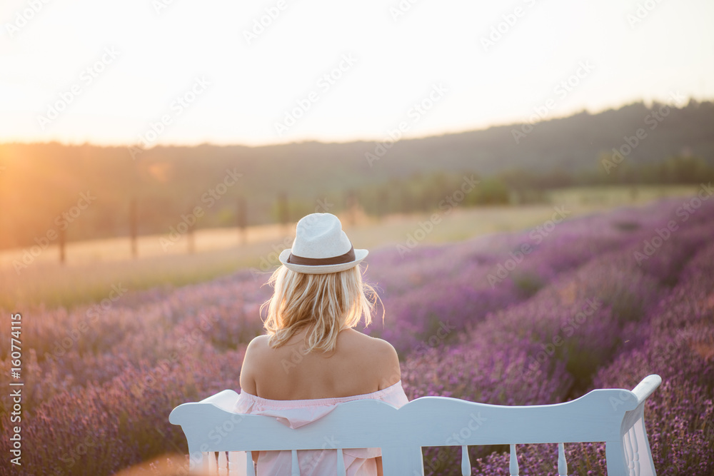 Wall mural Back view of beautiful long hair woman watching sunset at lavender field on a white vintage bench.