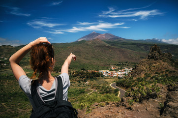 Fototapeta na wymiar Young woman with a backpack stands back to the camera and indicates by hand the peak of Teide volcano, Tenerife, Canary islands, Spain
