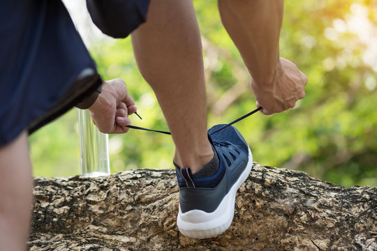 Cropped shot of young man runner tightening running shoe laces, getting ready for jogging exercise outdoors. Male jogger lacing his sneakers standing on forest path before morning run