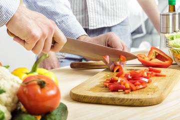 Hands of man chopped red bell pepper on board. Couple chopping vegetables in kitchen