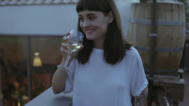 Shy unconfident, but pretty smart dressed young woman sips on spanish wine on open terrace with garden lights behind her, and party people casually dressed talk to each other