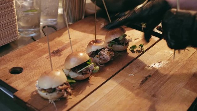 Asian fusion cuisine chef in authentic downtown bar prepares set of tiny mini burgers with pulled pork on artisan wooden tray, to serve to party crowd of millennials and young professionals