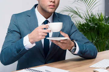 Cropped shot of stylish young businessman in suit holding cup of coffee and saucer