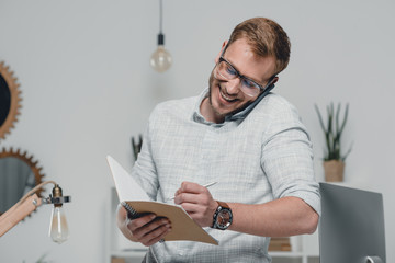 smiling caucasian businessman using smartphone and writing in diary in modern office