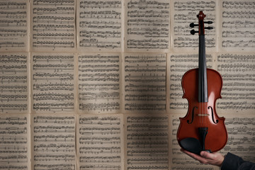 Vintage violin in man hands over the background of musical notes with lots of copyspace around. Arm of a male violin player rising out and holding an handcrafted violin, musical stringed instrument . © Gecko Studio