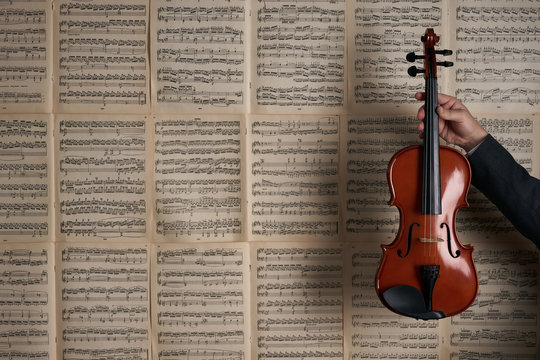 Vintage Violin In Man Hands Over The Background Of Musical Notes With Lots Of Copyspace Around. Arm Of A Male Violin Player Rising Out And Holding An Handcrafted Violin, Musical Stringed Instrument .