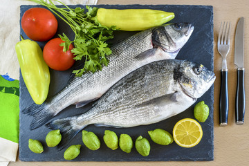 Two ready to cook raw fish with lemon and tomatoes on stone slate board.