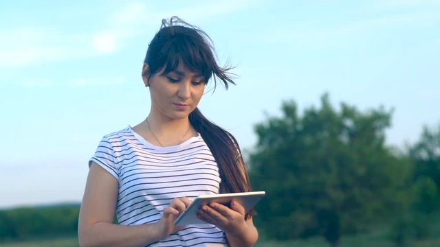 Young brunette girl with long hair writes a message on a tablet computer with a touch screen on a nature background