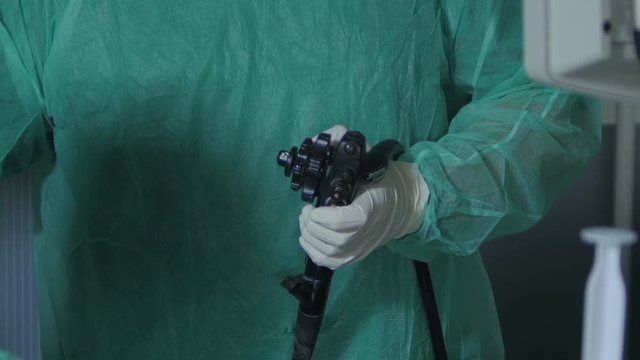 Tilt up from hands in protective gloves during using joystick to control micro camera for colonoscopy exam to portrait of middle aged male doctor gastroenterologist look on computer screen, indoors