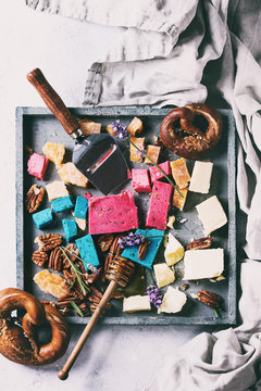 Variety of colorful holland cheese traditional soft, old, pink basil, blue lavender served with pecan nuts, honey, lavender flowers, pretzels bread on wood tray over gray texture background. Flat lay