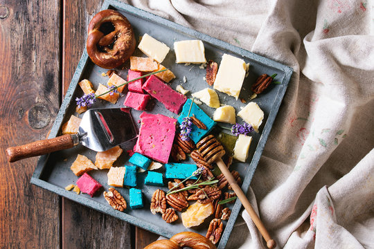 Variety of colorful holland cheese traditional soft, old, pink basil, blue lavender served with pecan nuts, honey, lavender flowers, pretzels bread on tray over wooden planks background. Flat lay