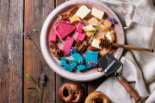 Variety of colorful holland cheese traditional soft, old, pink basil, blue lavender served with pecan nuts, honey, lavender flowers, pretzels bread on plate over wooden planks background. Flat lay