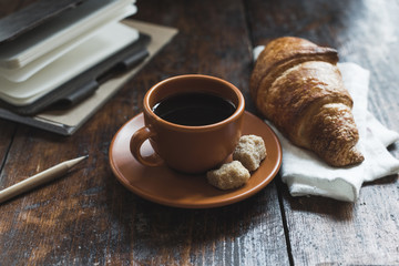 coffee with croissant on cloth and notebooks with pencil on wooden tabletop
