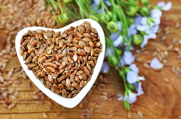 Flax seeds in heart-shaped bowl and blue linum plants, wooden background, selective focus