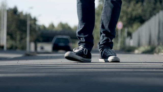 A man wearing a pair of sneakers taps his feet and stomps. Filmed from ground level and with a background of traffic in shallow focus.