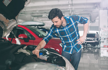 Perfect lines. The young dark-haired bearded man examining car at the dealership and making his choice. Horizontal portrait of a young guy at the car. He is thinking if he should buy it