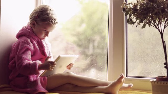 Cute little girl in pink bathrobe using tablet pc. Sitting on the window sill.