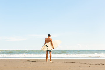 Handsome man stand with long surf surfing board wait on surf spot at sea ocean beach. View from back. White blank surfboard. Concept of sport, freedom, new modern life, generation Y.