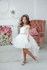 Beautiful little girl with curls and white Quinceanera dresses with a train on the background of the fireplace and chairs