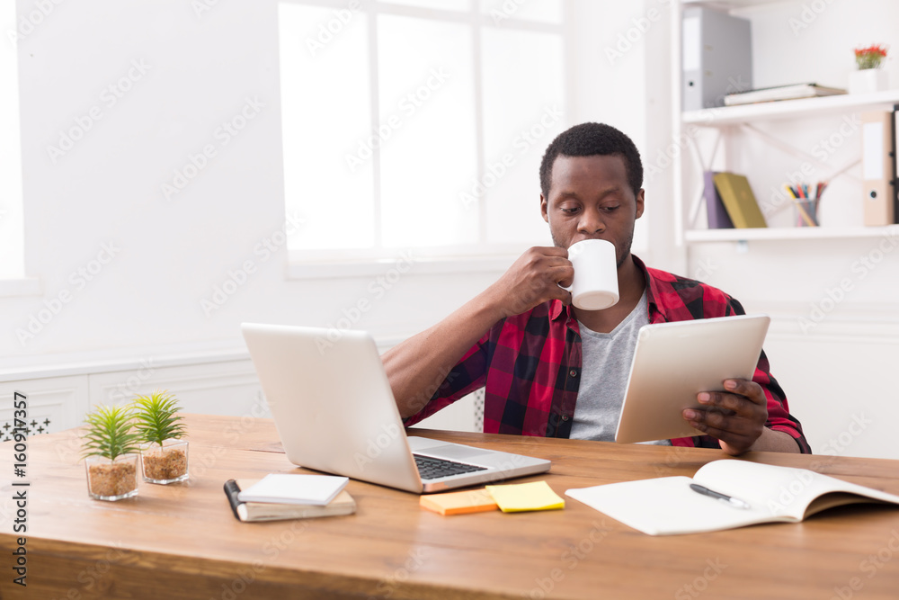 Wall mural Black businessman in casual office, reading news on tablet, drinking coffee