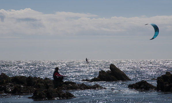 Girl with body board sitting on rocks looking out to parasailer