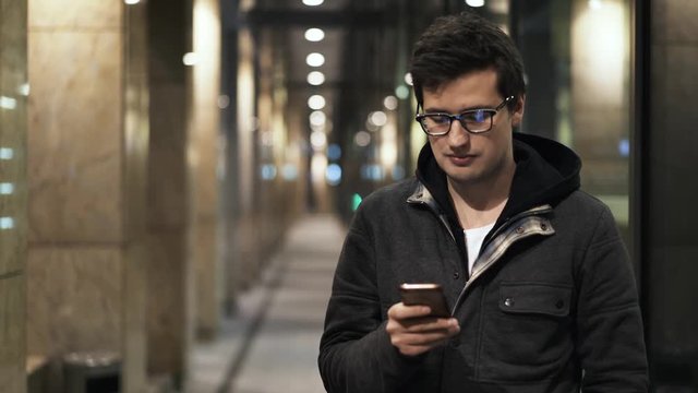 Portrait of a young man in glasses who is standing in a street and texting. Concept of modern communication. Locked down real time medium shot