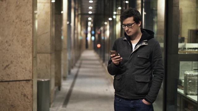 Young man wearing glasses and jeans with a jacket is standing in the street and texting. Locked down real time medium shot