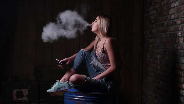 Young blonde woman smoking and vaping e-cigarette.Happy vaping young white blonde girl. smoking fruit flavored e-liquid or e-juice with vaporizer device or e-cig. Modern gadget for smokers