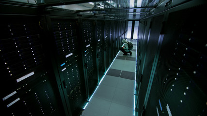 Security Camera view of a Hooded Hacker in Data Center, With His Laptop He Connects to One of The Rack Servers, Commits Crime.